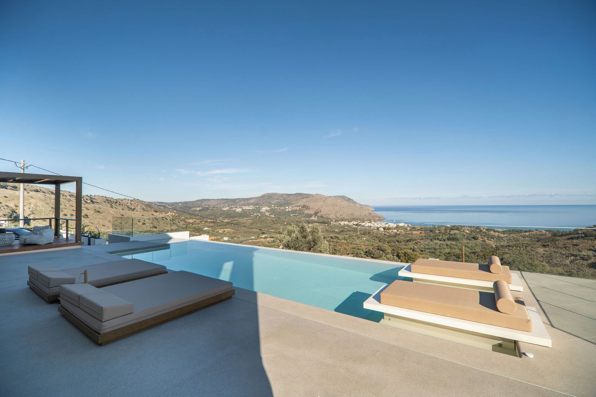 A rooftop infinity pool with lounge chairs, a pergola, and a scenic view of the sea and hills.
