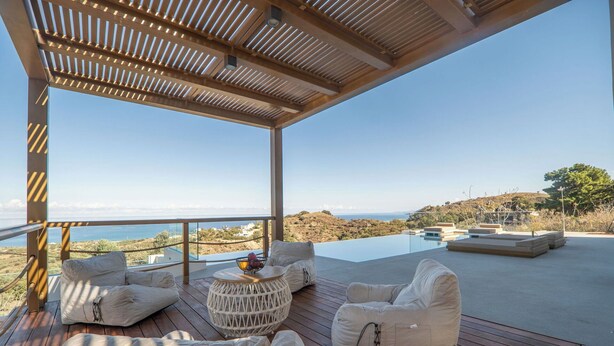 A modern outdoor lounge area with a view of the sea, featuring a wooden deck, white cushioned seating, and a small round table. - Lofos Orizon Villa - Villa with pool in Crete (Mathés)