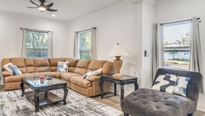Ein Wohnzimmer mit einem Ecksofa, einem Glastisch, einem Beistelltisch mit Lampe und einem gepolsterten Hocker.