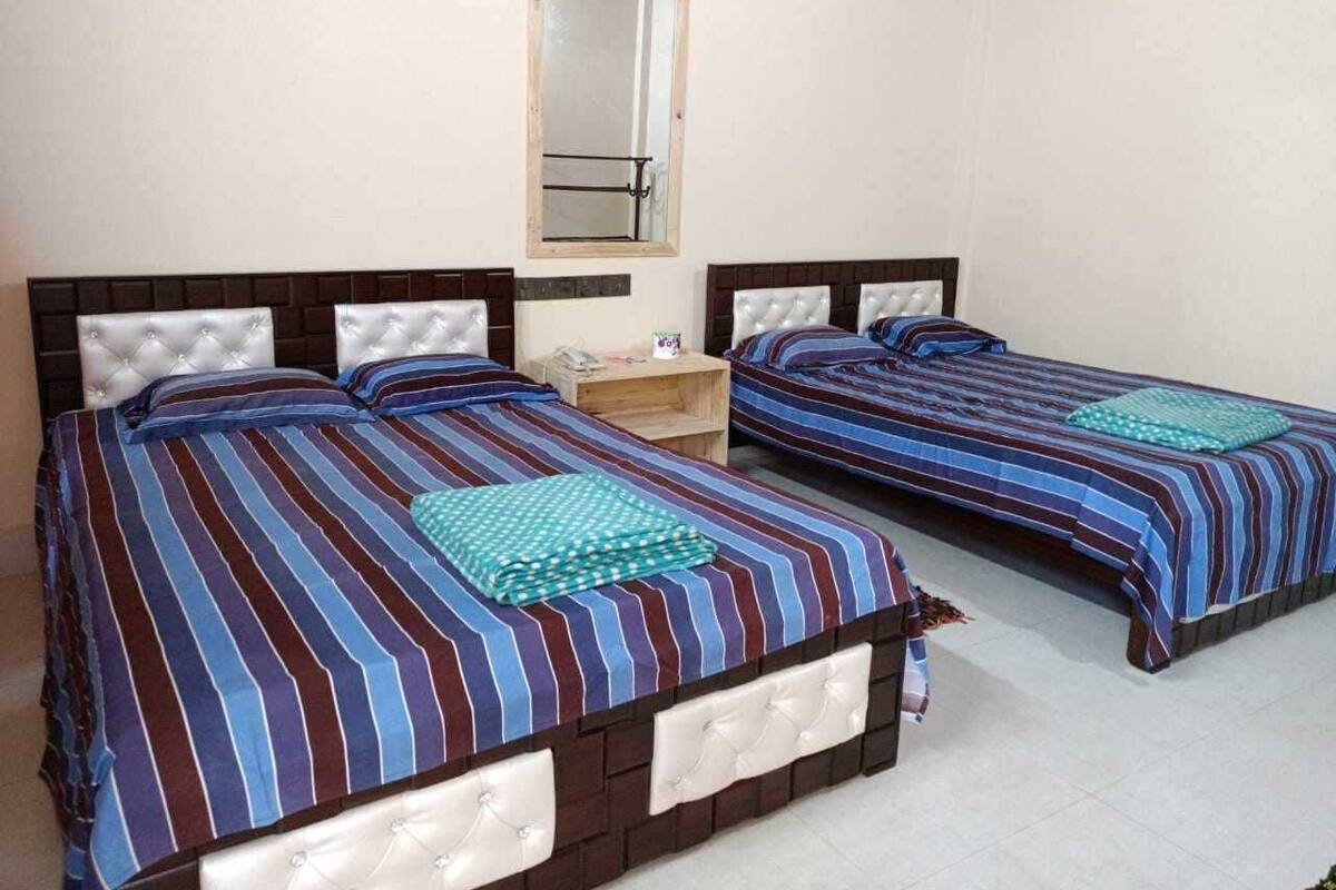 Ein Zimmer mit zwei Betten, jeweils mit gestreifter Bettwäsche und einem gepolsterten Kopfteil. Zwischen den Betten steht ein Nachttisch aus Holz, und über dem linken Bett befindet sich ein Spiegel an der Wand.