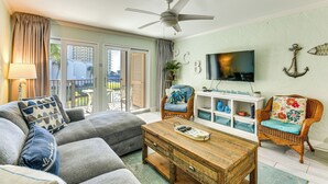 A living room with a ceiling fan, a TV, wicker furniture, and a view to the outside. - Walk to Shore + Restaurants: Resort Retreat in PCB (Panama City Beach)