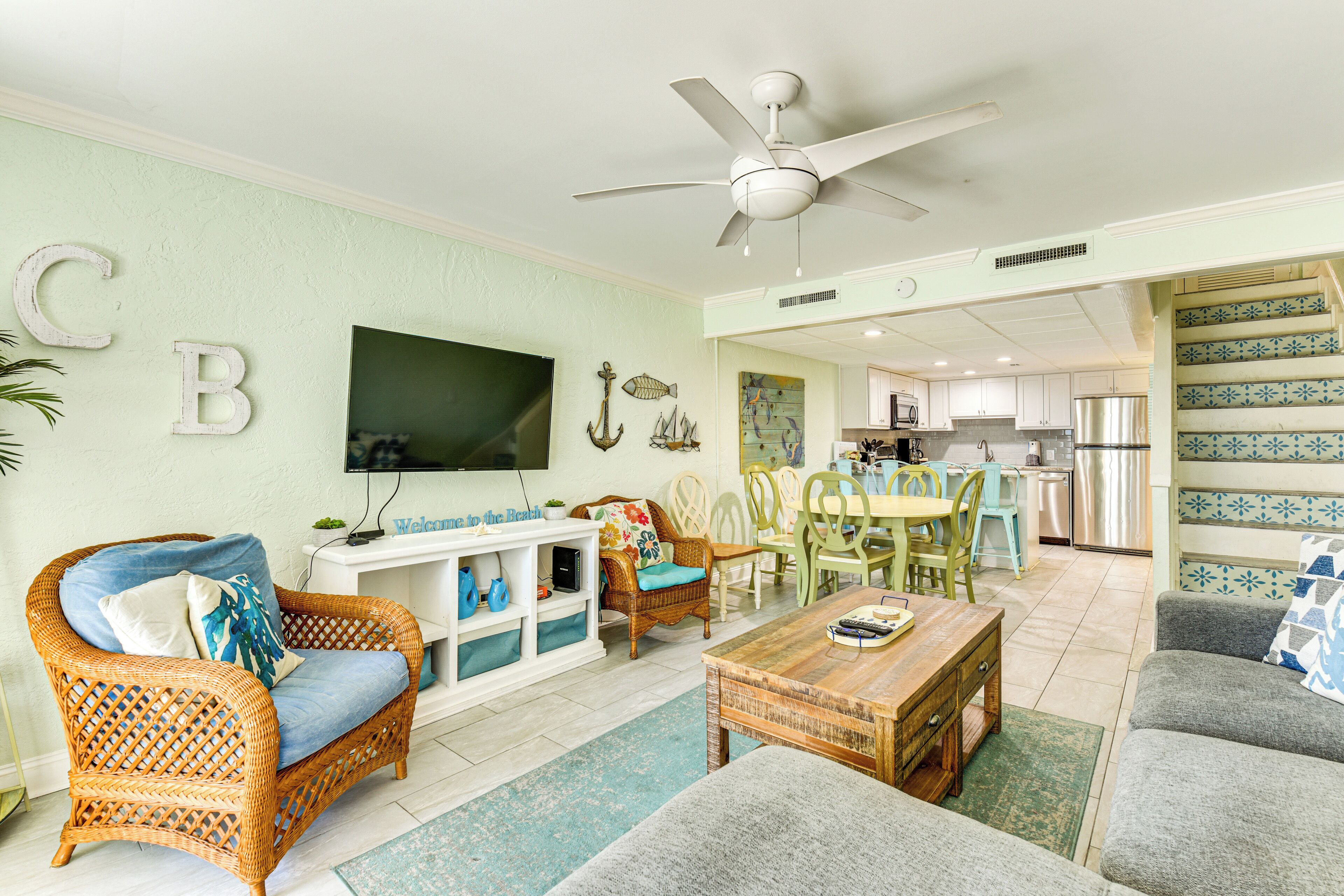 A living room with wicker furniture, a flat-screen TV, and a kitchen in the background.