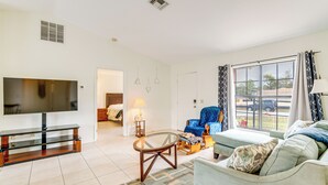 A living room with a flat-screen TV, a glass-top coffee table, a blue armchair, a sofa, and a large window with curtains.