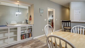Un comedor con una mesa de madera y sillas blancas, una estantería con libros y un espejo sobre la mesa.