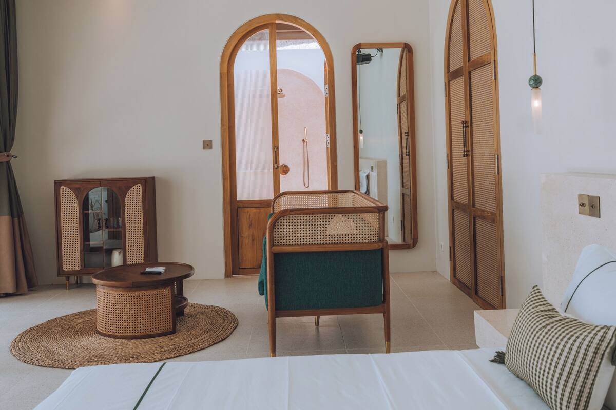 A modern hotel room with a large bed, a wooden wardrobe, a wicker chair, a small round table, and a large mirror.