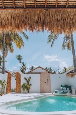 A pool area with a thatched roof, palm trees, and a white wall with wooden doors. - Lumora Boutique Villas (Gili Trawangan)