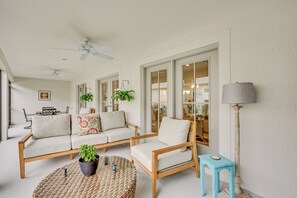 A living room with a sofa, armchair, and a small table with a plant. There are ceiling fans, a lamp, and a view of the kitchen through glass doors. - Beautiful Garden Villa with Golf Cart Option (Sumterville)