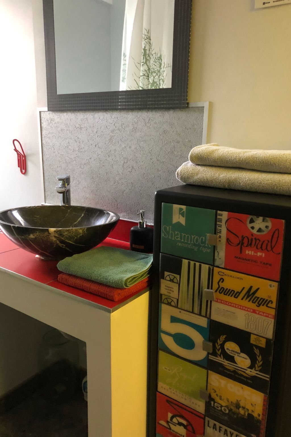 A bathroom with a large bowl sink, a mirror, and a refrigerator with various tapes and towels.