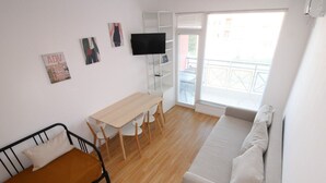 Um espaço compacto com cama, sofá, mesa de jantar com cadeiras, TV e varanda com guarda-corpo.