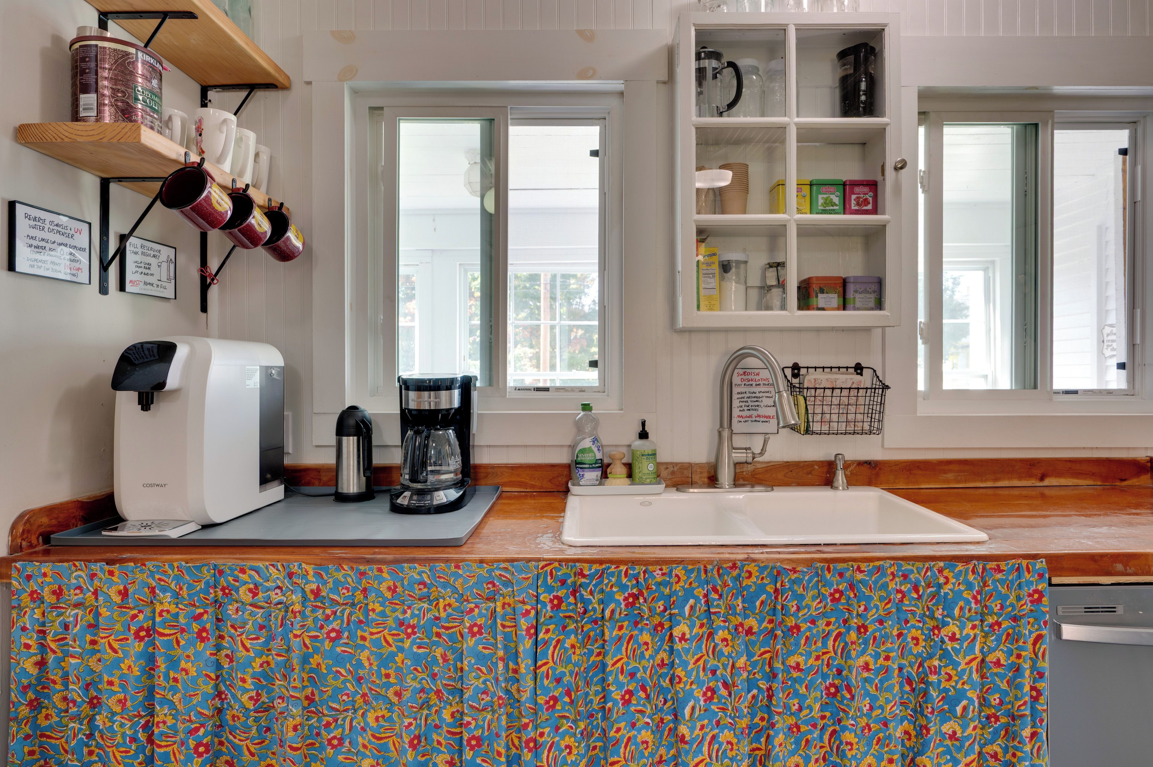 A kitchen with a coffee maker, a kettle, and a sink. There are shelves with jars and a window with a view.