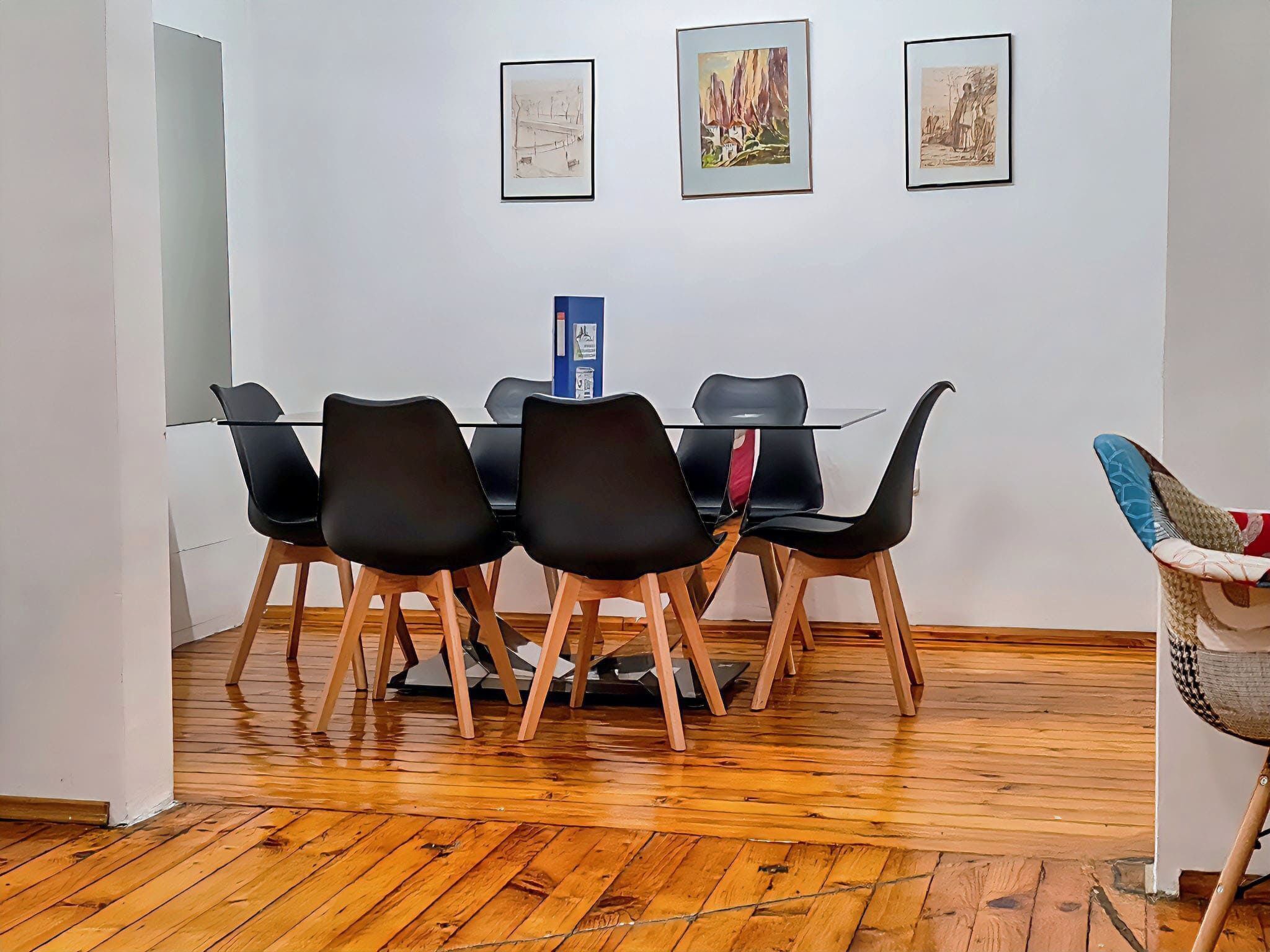 A dining area with a glass table and black chairs, wooden flooring, and three framed artworks on the wall.