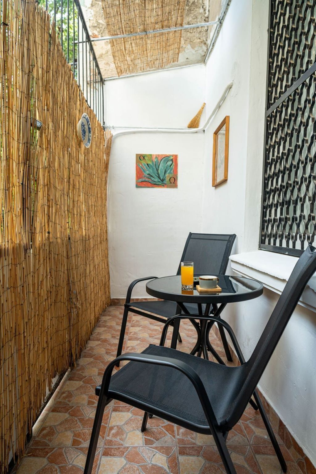Un petit patio clos avec une table ronde et deux chaises, un verre de jus d’orange et un tableau encadré accroché au mur.