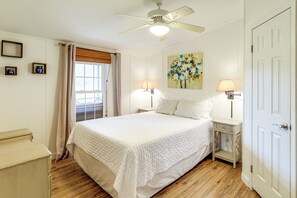 A bedroom with a large bed, two bedside tables with lamps, a ceiling fan, a window with curtains, and a door.