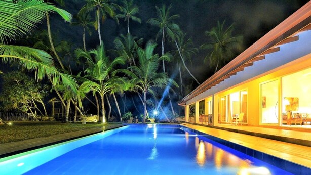 Une piscine la nuit, avec des palmiers en arrière-plan et un bâtiment aux grandes fenêtres sur la droite.
