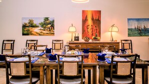 Un coin repas avec une longue table dressée pour six personnes, entourée de chaises en bois dont les dossiers sont en osier tressé. Deux lampes murales sont fixées de part et d’autre de la table.