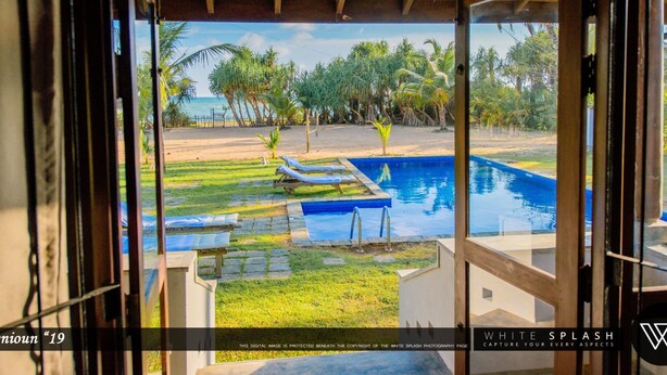 A view from a window of a pool area with a clear sky, palm trees, and a beach in the background.