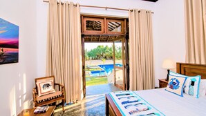 A hotel room with a bed, a wooden headboard, a nightstand, a lamp, a picture frame, a window with curtains, and a view of a swimming pool and greenery outside.