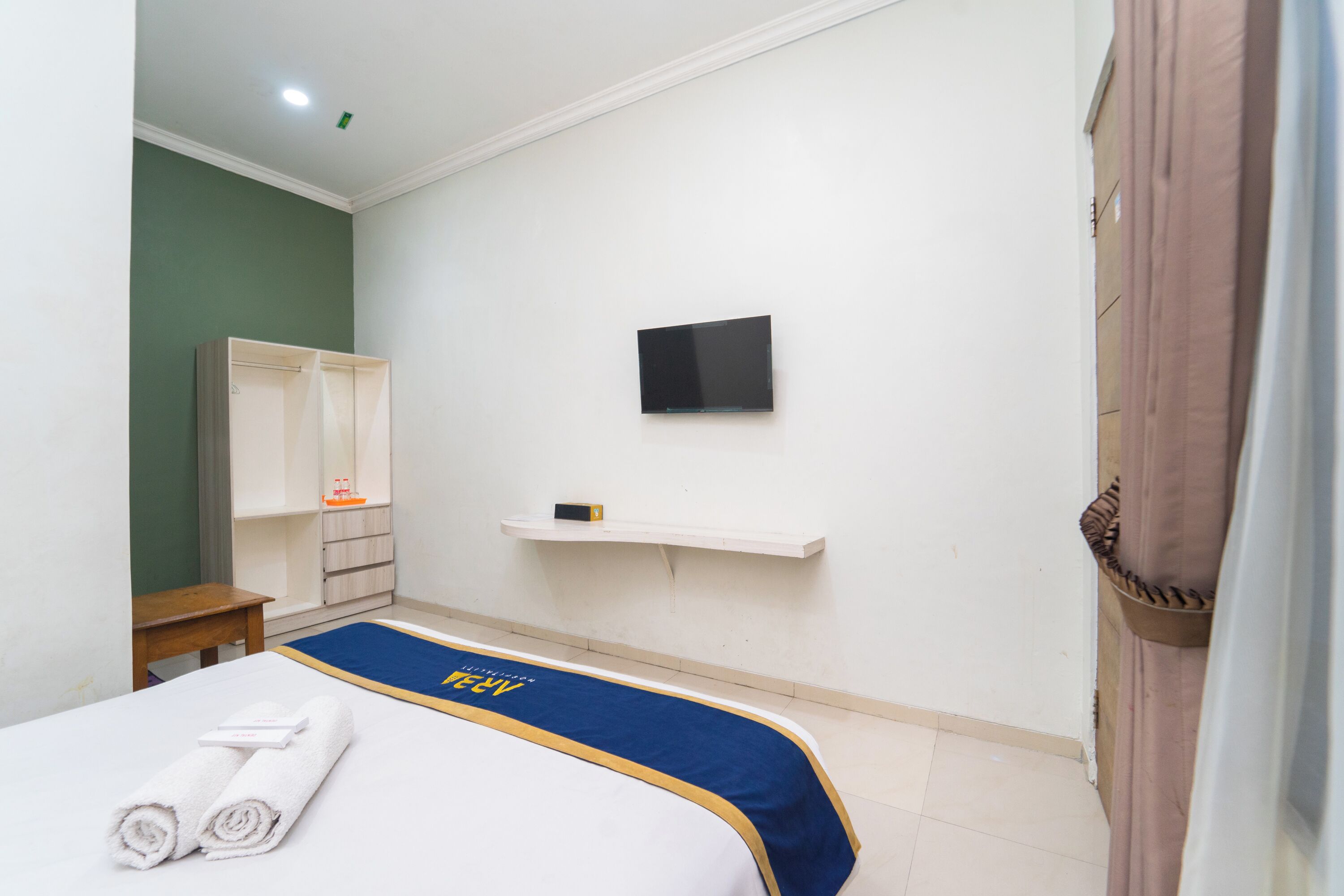 Une chambre d’hôtel avec un lit, un téléviseur fixé au mur, une table de chevet en bois et une étagère contenant des articles de toilette.