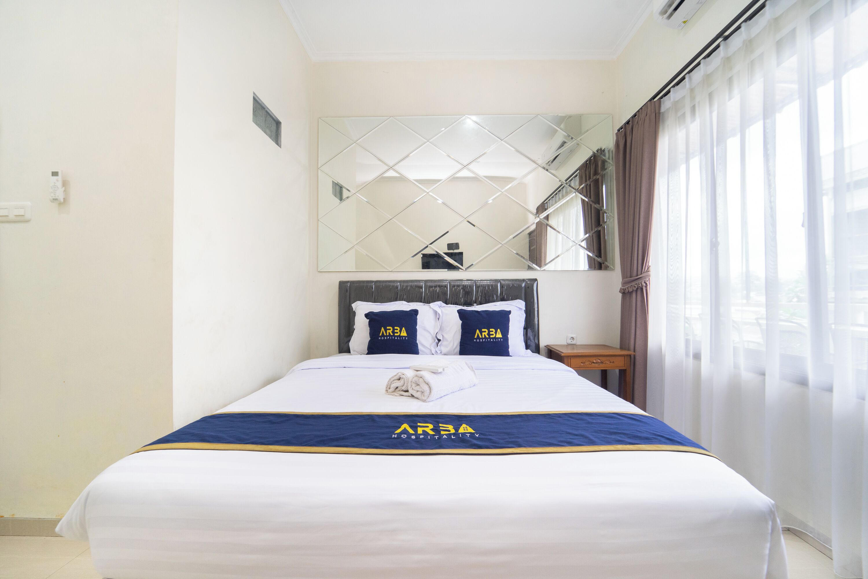 A hotel room with a neatly made bed, a large mirror above the bed, and a view of a staircase.