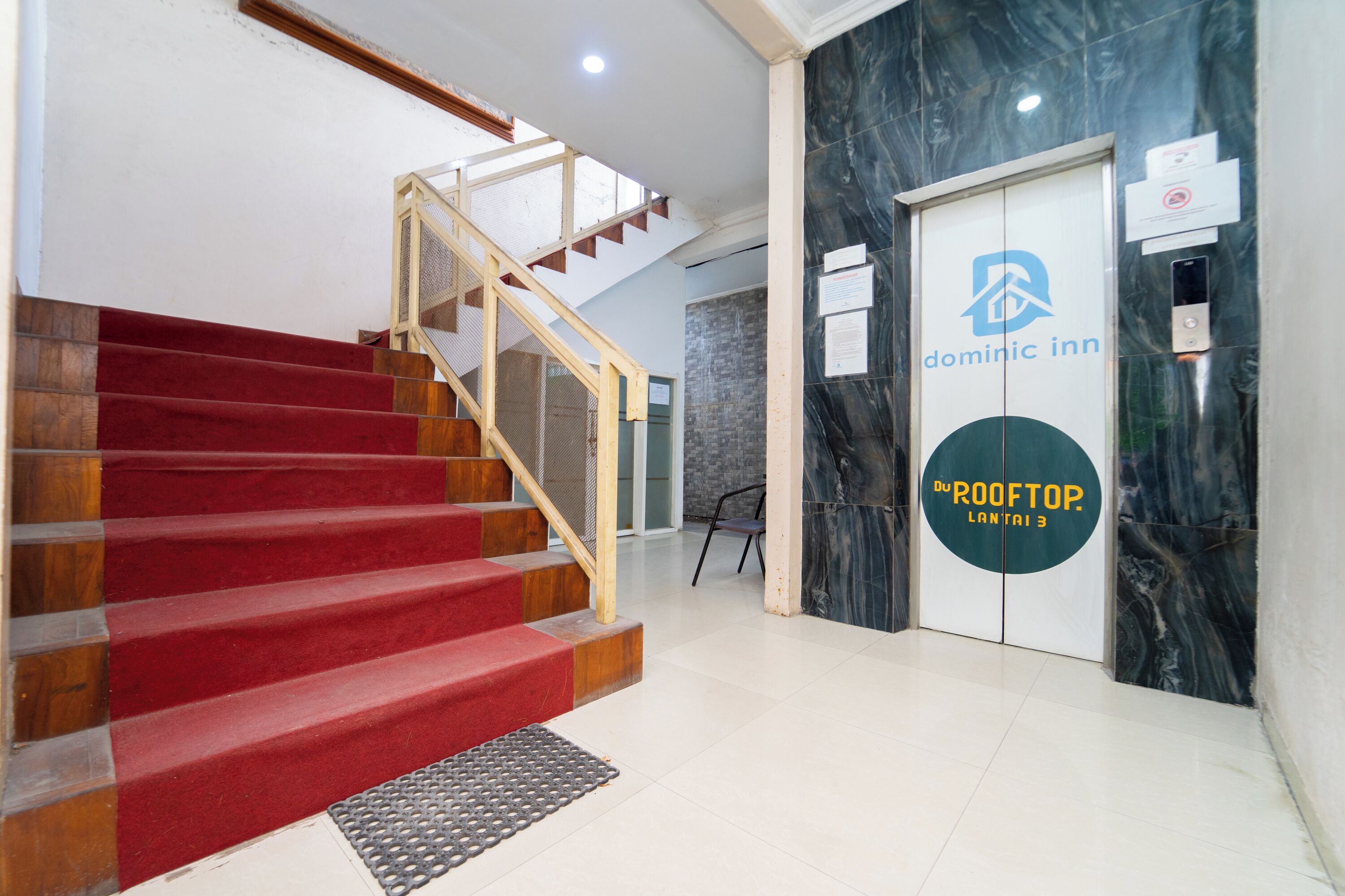 A hotel lobby with a staircase leading to an upper floor, a reception desk, and a sign for 'Dominic Inn' and 'Du ROOFTOP LANTAI 3'.