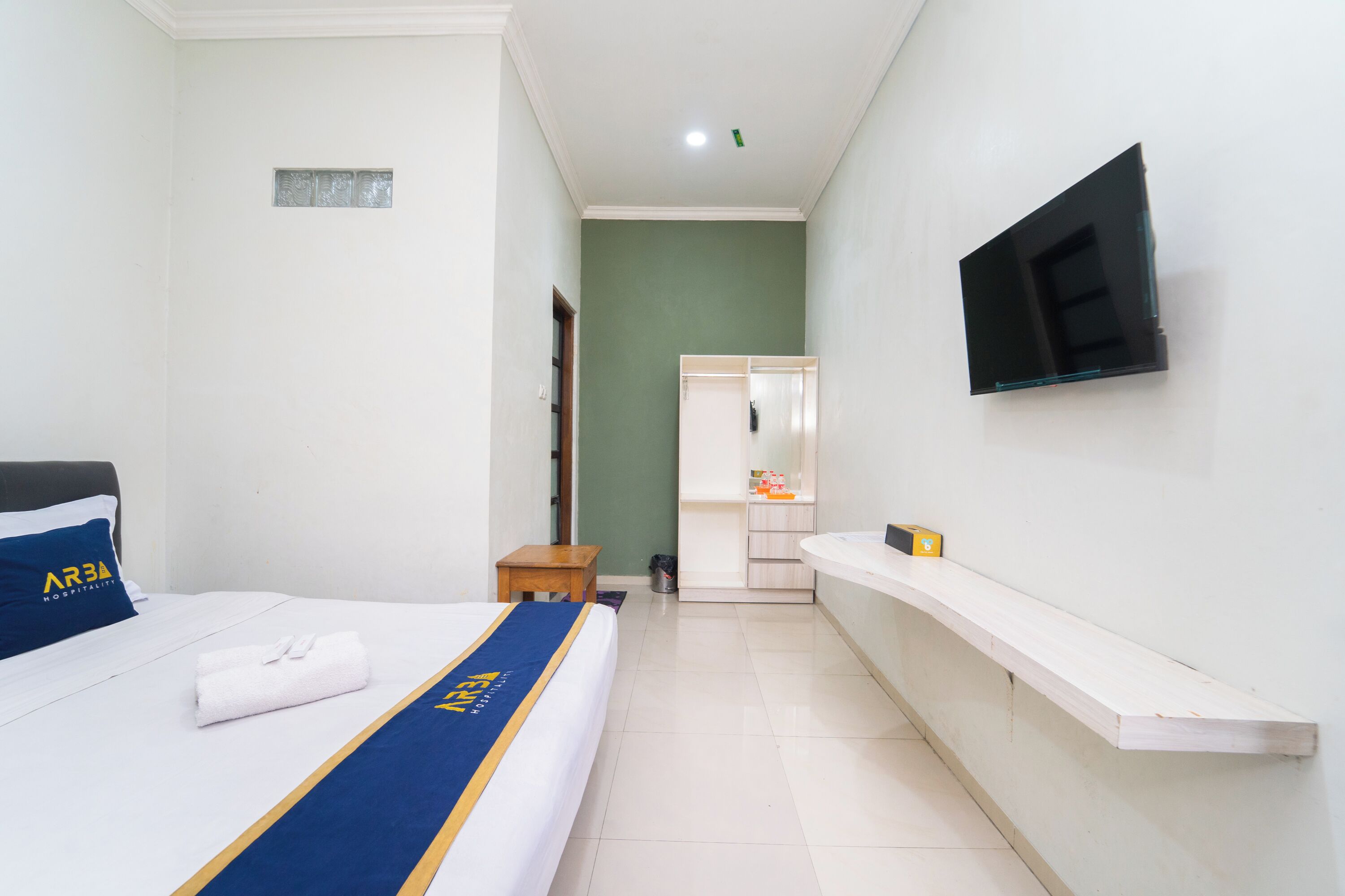 A hotel room with a bed, a TV mounted on the wall, a small table, and a bench.