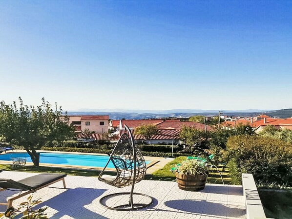 A poolside area with a hanging chair, lounge chairs, and a view of a residential area with red-roofed buildings.