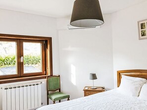 Un dormitorio con cama, sillón, mesita de noche, lámpara, ventana y radiador.