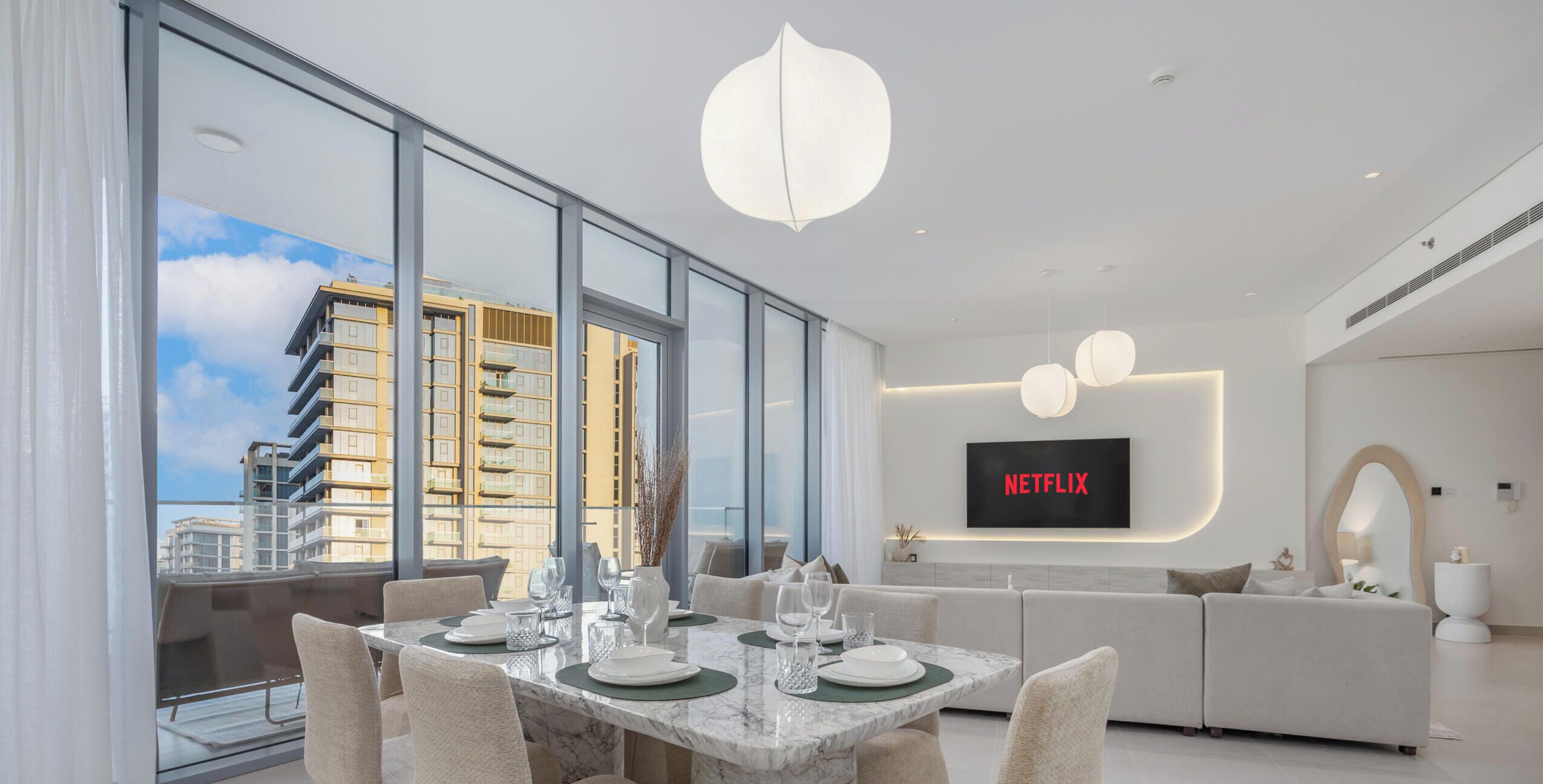 A modern living room with a dining area, large windows, a flat-screen TV, and a sofa.