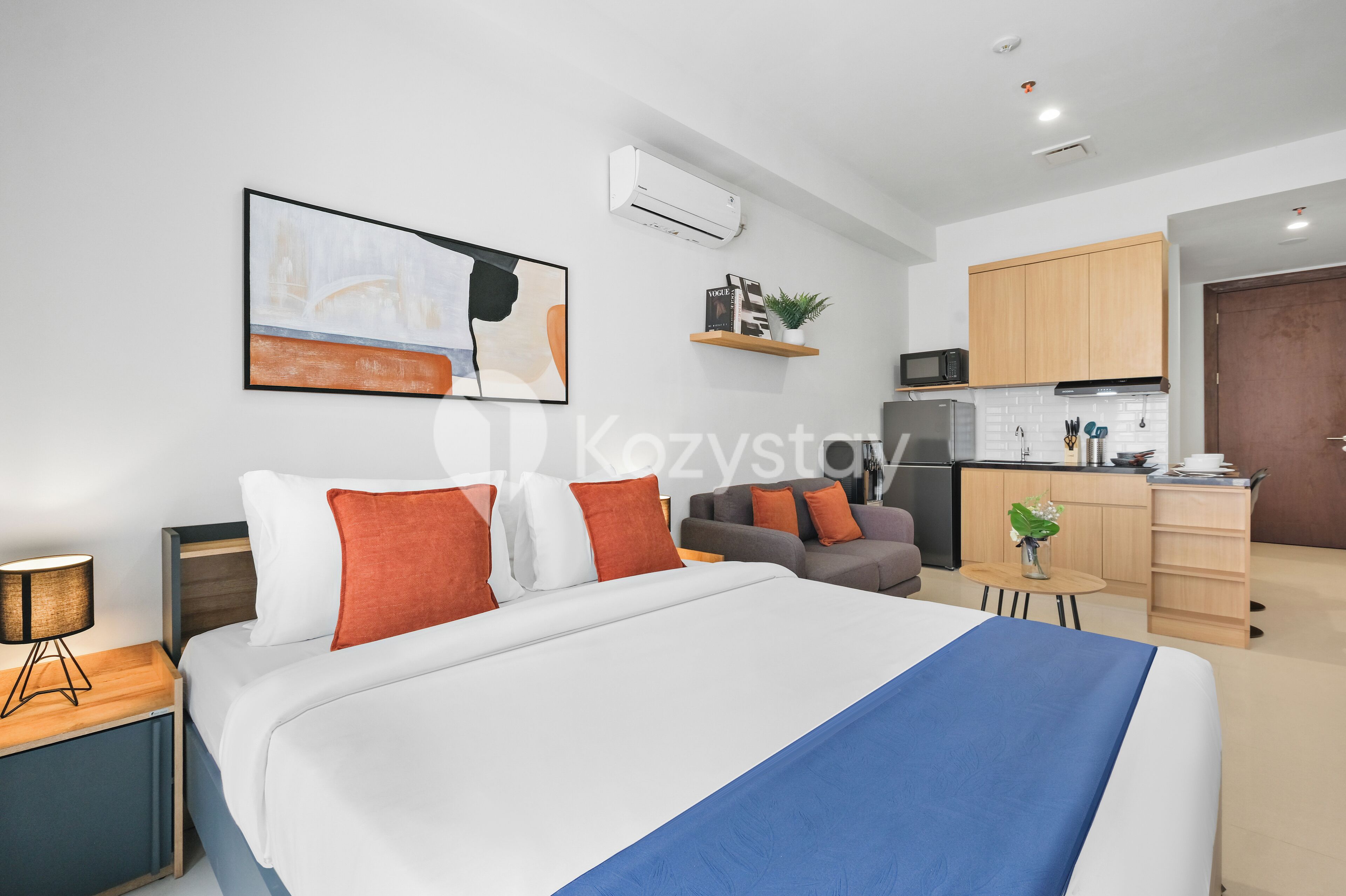A modern hotel room with a large bed, a desk, a sofa, a kitchen area, and a dining table.