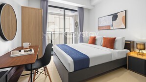 A hotel room with a bed, a desk with a chair, a bedside table with a lamp, a window with curtains, and a wall-mounted artwork. - Hesrin by Kozystay - Sentul (Bogor)