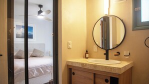 A bathroom with a round mirror, a sink, and a bed visible through an open door. - Cozy 2-bedroom house in El Tesoro with AC (El Tesoro)