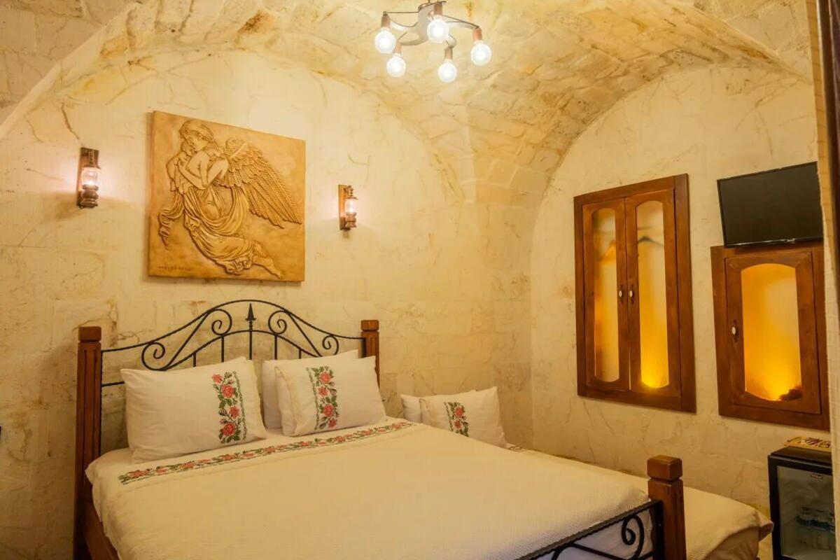 Quarto com parede de pedra, cama com almofadas florais, pintura de um anjo e armários de madeira com televisão.