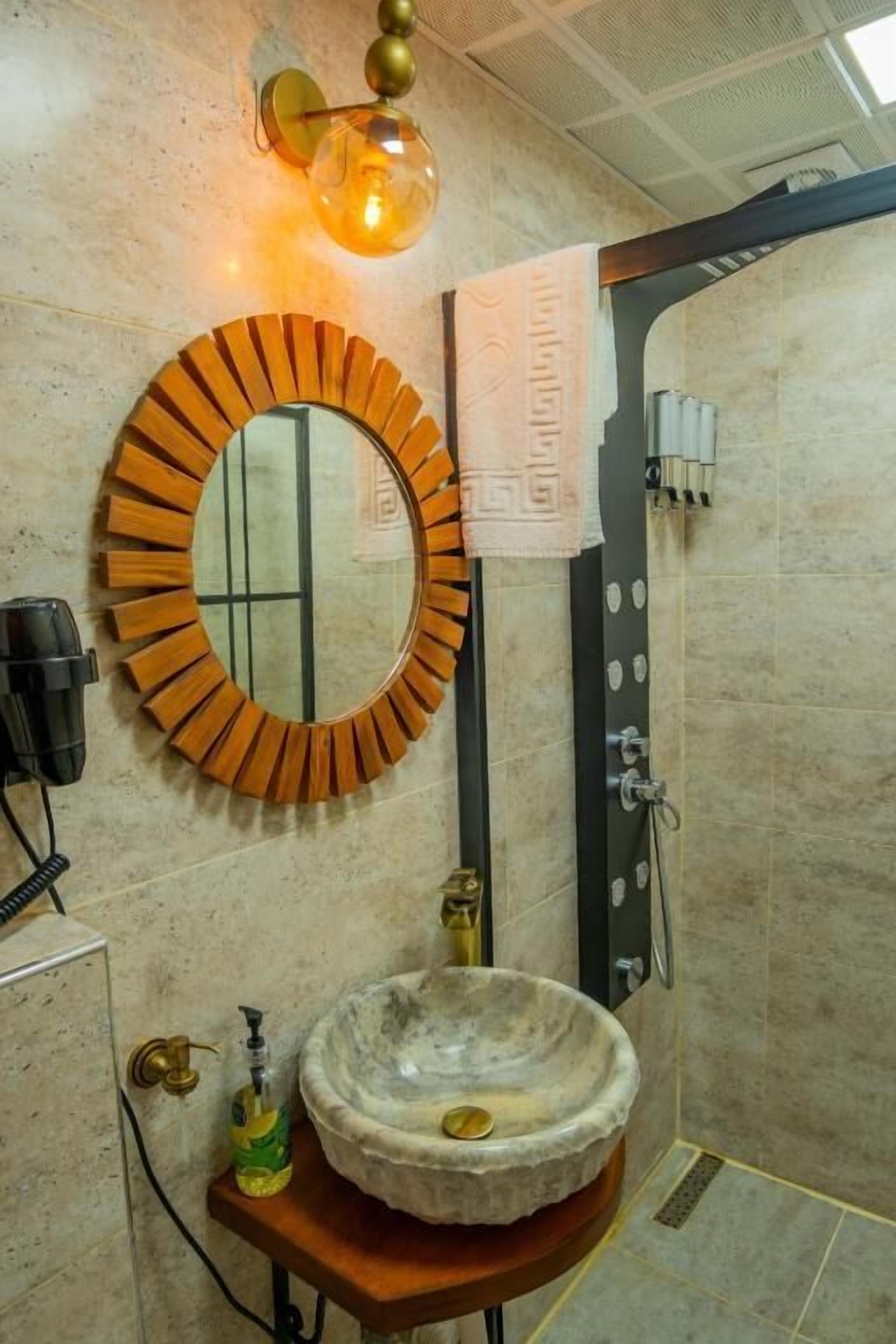 A bathroom with a round mirror, a stone sink, a towel hanging on the wall, and a shower area.