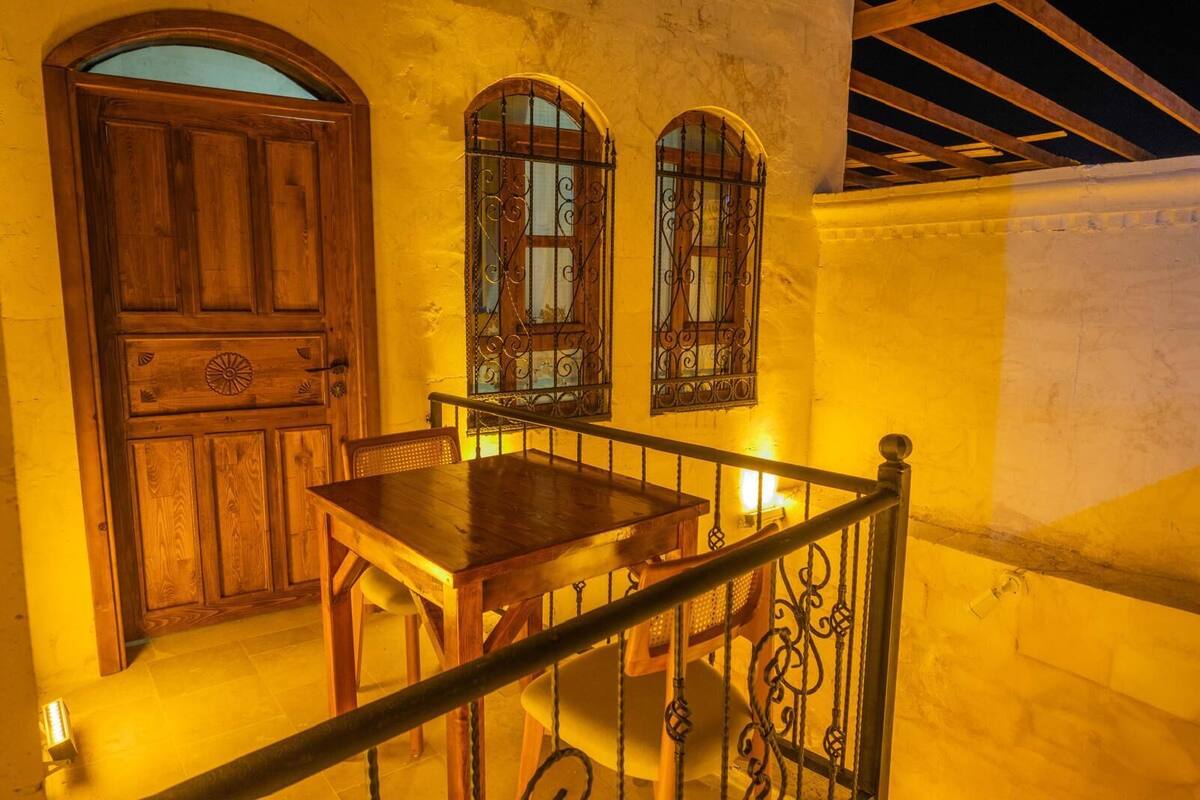 Conjunto de mesa e cadeiras de madeira, com uma parede amarela e duas janelas em arco com grades de metal.