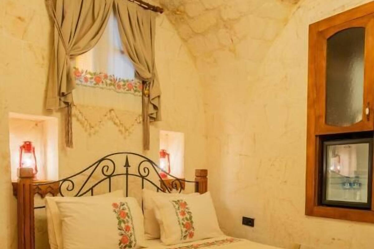 Um quarto com teto de pedra, estrutura de cama em madeira, tapete vermelho estampado e janela com cortinas.
