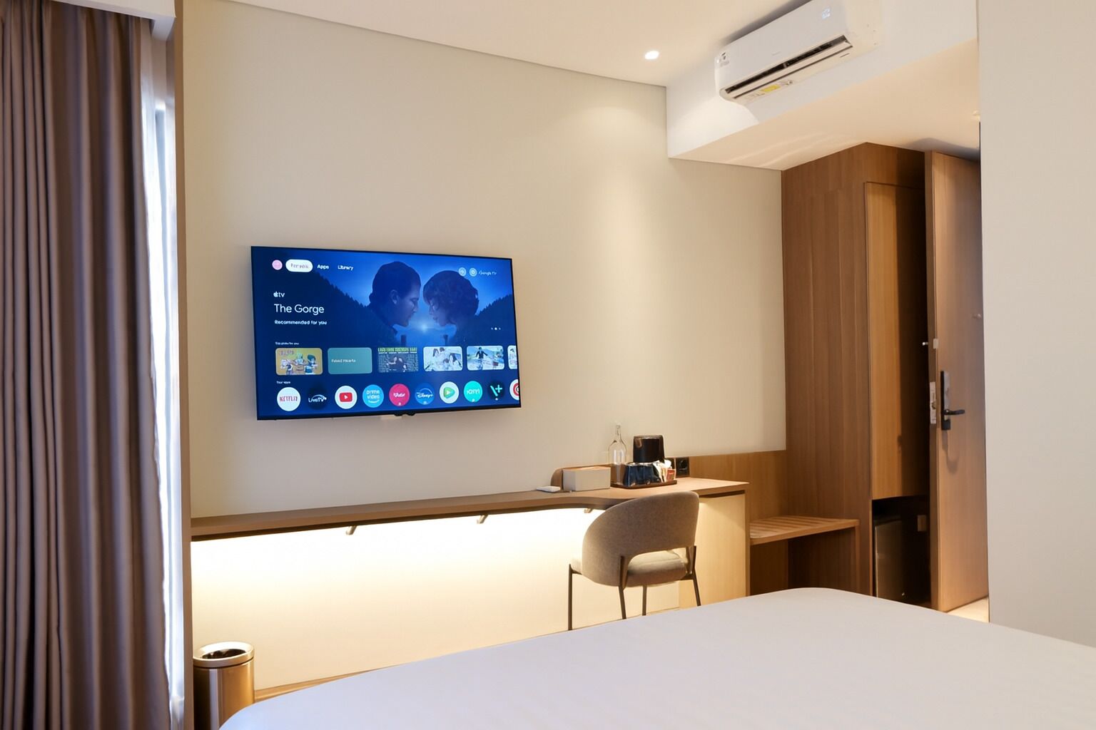A modern hotel room with a flat-screen TV mounted on the wall displaying a TV guide. There is a desk with a chair, a bedside table, and a wardrobe.