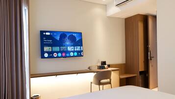 A modern hotel room with a flat-screen TV mounted on the wall displaying a TV guide. There is a desk with a chair, a bedside table, and a wardrobe.