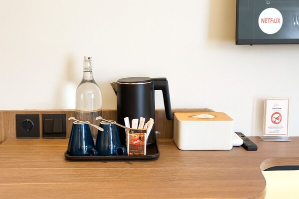 A hotel room with a wooden desk, a coffee maker, a glass bottle, a black kettle, a box of tissues, a remote control, and a framed sign. - The Artha - Bandung (Bandung)