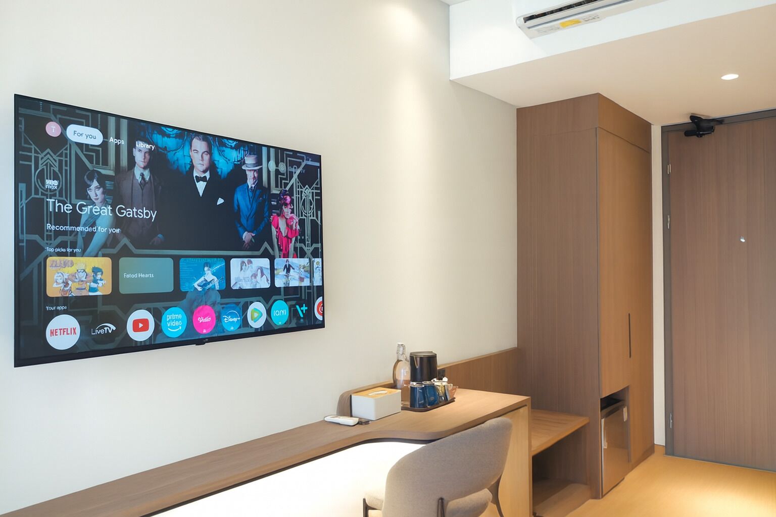 A modern hotel room with a large wall-mounted TV displaying a movie recommendation, a wooden desk with a chair, and a wooden wardrobe.