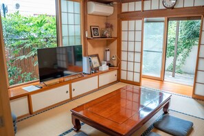 Ein traditioneller japanischer Raum mit Tatami-Bodenbelag, einem niedrigen Tisch, einem Fernseher und einer Schiebetür, die in einen Garten führt.
