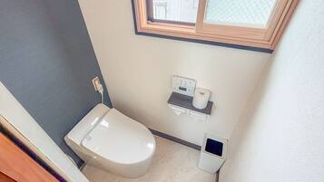 A small, compact bathroom with a toilet, a hand dryer, a paper towel dispenser, and a window above the toilet.