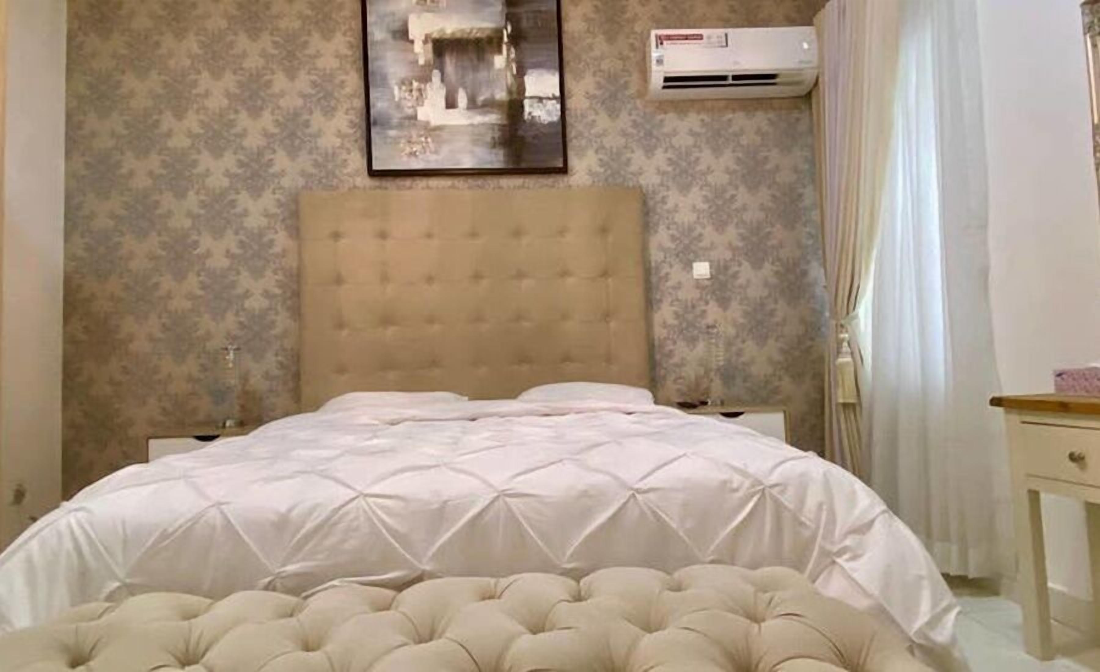 A bedroom with a tufted headboard, a bed with a white duvet, a nightstand, an air conditioning unit, and a framed picture on the wall.