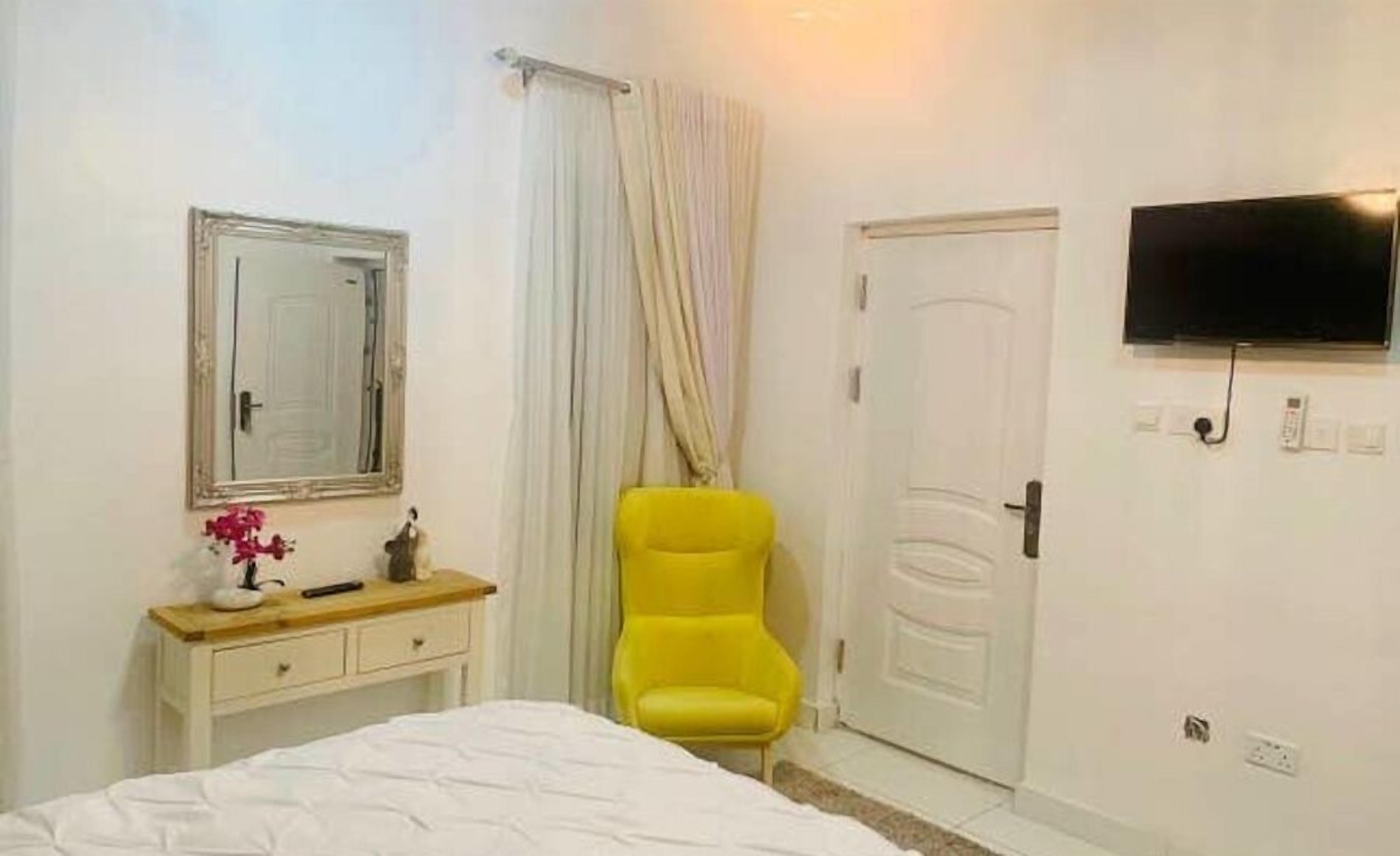 Una camera da letto con un letto, un comodino con lampada, uno specchio, una sedia gialla, una porta bianca e una TV montata a parete.
