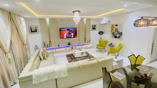 Uma sala de estar moderna com um sofá bege, poltronas amarelas, uma mesa de centro de vidro e uma TV fixada na parede.