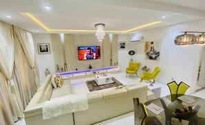 Una sala de estar moderna con un sofá color beige, sillas amarillas, una mesa de centro de vidrio y un televisor montado en la pared.