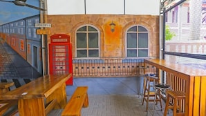 Zona de bar con bancos de madera, un teléfono rojo y una pared de ladrillo con ventanales arqueados.