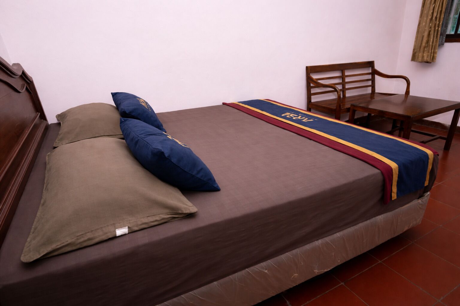 A single bed with a brown mattress, two pillows, a blue blanket, and a red and blue striped bedspread.