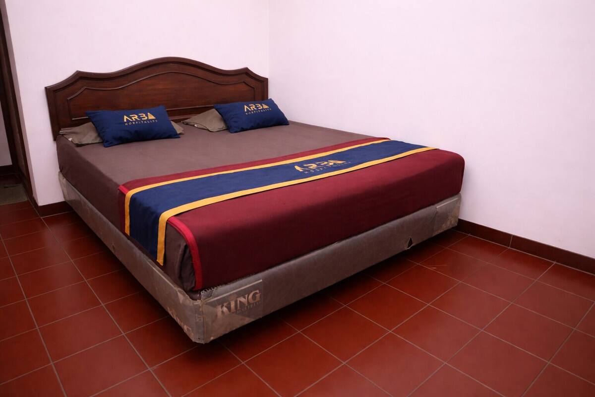 Un llit individual amb capçalera de fusta, un llençol de llit vermell i blau, i dos coixins amb la inscripció "AR3A HOSPITALITY".