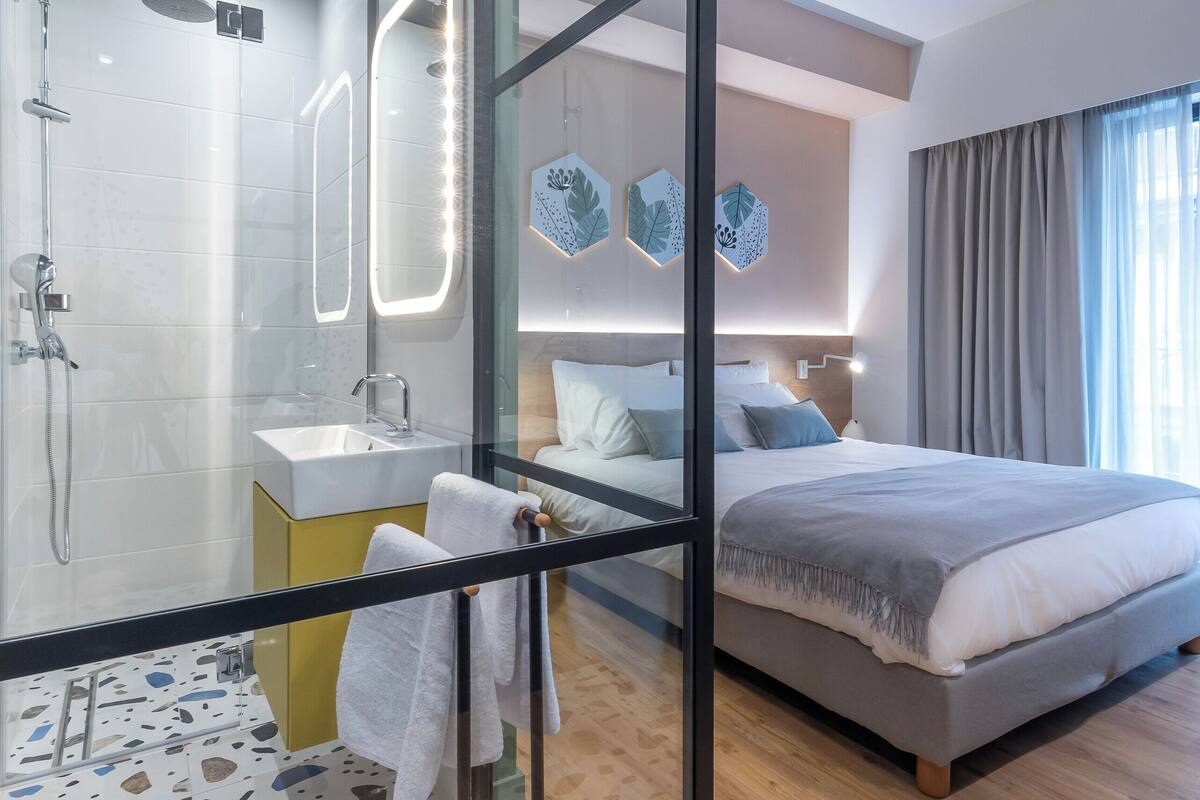 A modern hotel room with a glass shower enclosure, a bedside table with a sink, a bed with a gray blanket, and a large window with curtains.