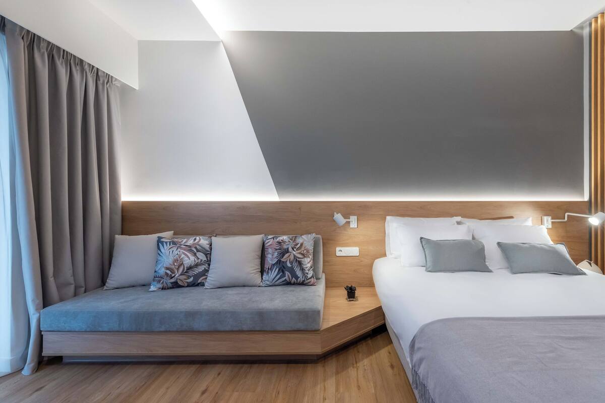 A modern hotel room with a sofa bed, a separate bed, and a wooden headboard.